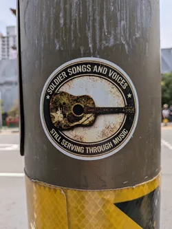 Street sticker A circular sticker with a distressed, vintage aesthetic. It features a central acoustic guitar decorated with a camouflage pattern and a star. The guitar is set against a rusty, off-white background, enclosed by a black circular border containing the text 'SOLDIER SONGS AND VOICES' at the top and 'STILL SERVING THROUGH MUSIC' at the bottom, separated by two small stars.