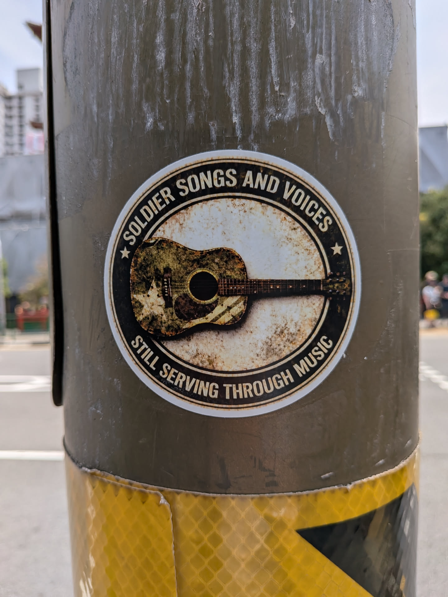 A circular sticker with a distressed, vintage aesthetic. It features a central acoustic guitar decorated with a camouflage pattern and a star. The guitar is set against a rusty, off-white background, enclosed by a black circular border containing the text 'SOLDIER SONGS AND VOICES' at the top and 'STILL SERVING THROUGH MUSIC' at the bottom, separated by two small stars.