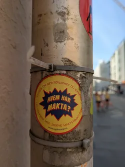 Street sticker A circular yellow sticker with a prominent jagged blue starburst in the center. Inside the starburst, the question "HVEM HAR MAKTA?" (Who has the power?) is written in bold red letters. Encircling the starburst, along the yellow border, is text in black that reads "KAMPEN OM RETTFERDIGHET OG BÆREKRAFT I ET SAMFUNN MED SKEVE MAKTSTRUKTURER" (The fight for justice and sustainability in a society with skewed power structures). At the very bottom, the website "www.spireorg.no" is displayed. The sticker is attached to a metallic pole, showing signs of wear and tear, including fading and some dirt.
