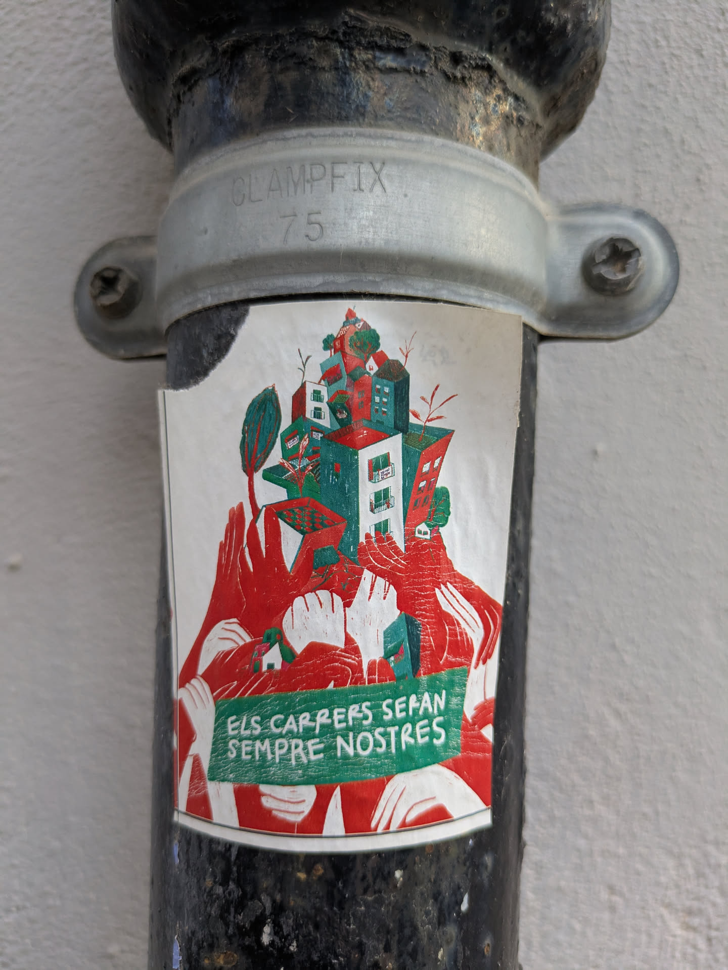 The sticker features an illustration of a cityscape with buildings and trees, all rendered in red, green, and white. The cityscape appears to be supported by many hands. At the bottom, there's text in a banner with a green background. The text is in Catalan.