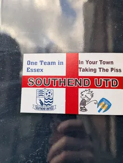The sticker features a St. George's Cross (England flag) as its background. Prominently displayed across the center is the text 'SOUTHEND UTD' in large black letters with a white outline, set against the red horizontal stripe of the cross. In the top left quadrant, blue text reads 'One Team in Essex', while the top right quadrant contains dark red text stating 'In Your Town Taking The Piss'. The bottom left quadrant showcases the official crest of Southend United, featuring a shield with three seaxes, a football, a shrimp, and waves, with 'SOUTHEND UNITED' written on a banner below. The bottom right quadrant depicts a cartoon character, strongly resembling Calvin from Calvin and Hobbes, in a urinating pose, with the stream directed at a shield-shaped crest for 'COLCHESTER UNITED FC', which features two golden eagles on a blue and yellow background.