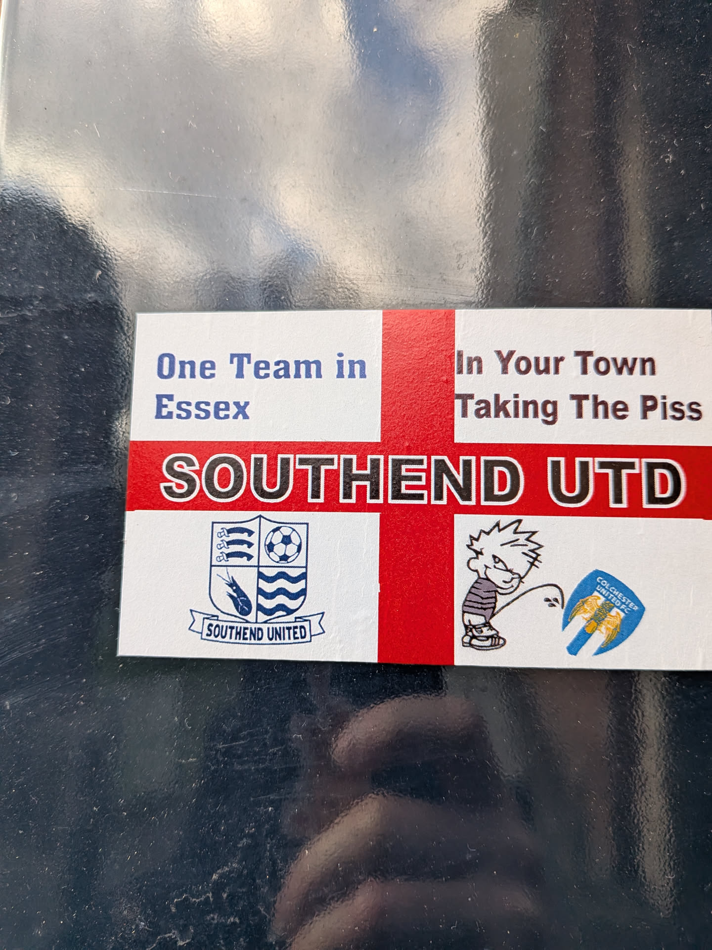 The sticker features a St. George's Cross (England flag) as its background. Prominently displayed across the center is the text 'SOUTHEND UTD' in large black letters with a white outline, set against the red horizontal stripe of the cross. In the top left quadrant, blue text reads 'One Team in Essex', while the top right quadrant contains dark red text stating 'In Your Town Taking The Piss'. The bottom left quadrant showcases the official crest of Southend United, featuring a shield with three seaxes, a football, a shrimp, and waves, with 'SOUTHEND UNITED' written on a banner below. The bottom right quadrant depicts a cartoon character, strongly resembling Calvin from Calvin and Hobbes, in a urinating pose, with the stream directed at a shield-shaped crest for 'COLCHESTER UNITED FC', which features two golden eagles on a blue and yellow background.