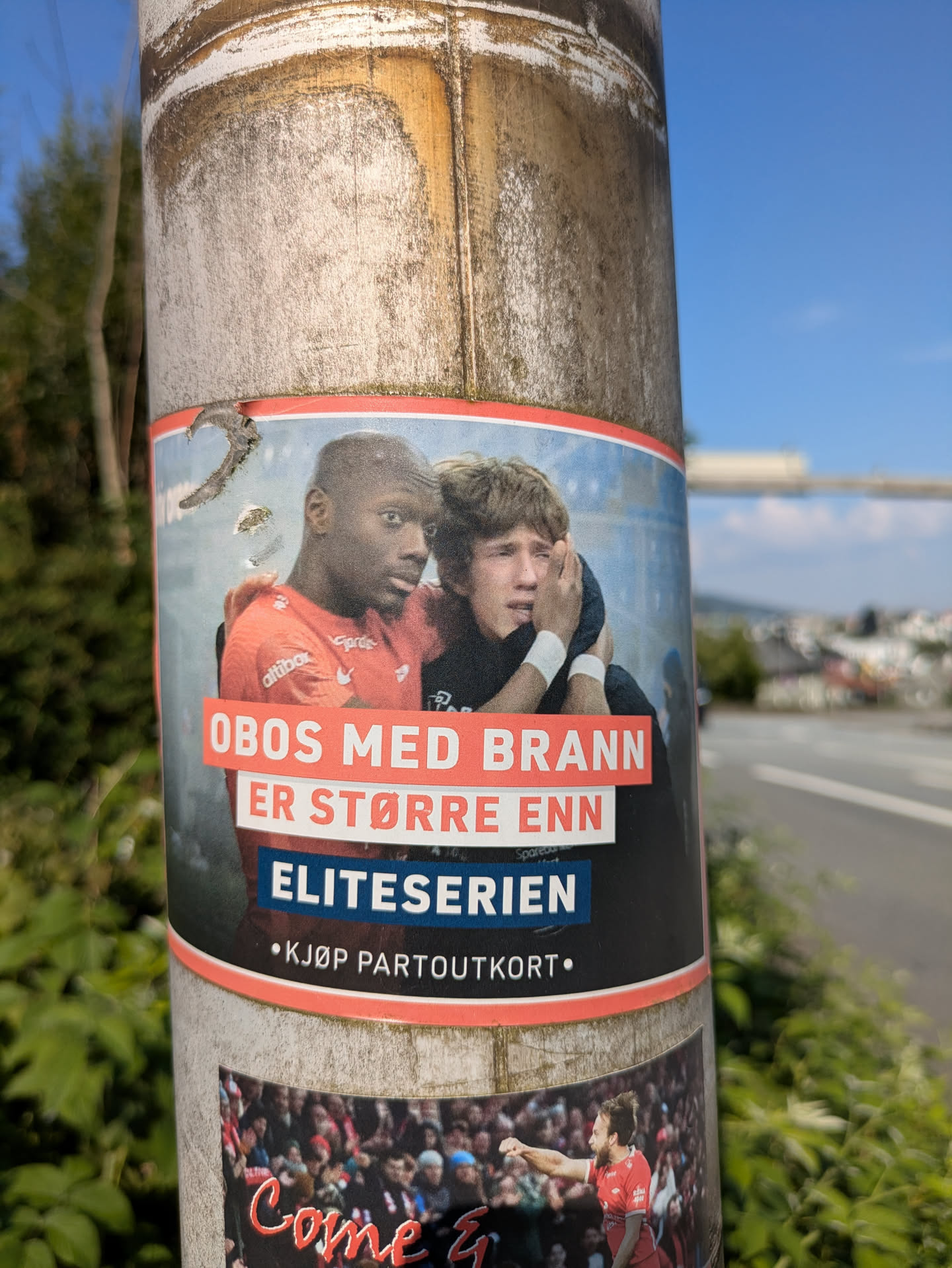 The sticker prominently displays two male football players, one with dark skin wearing a red jersey (likely SK Brann's colors) and another with light skin and curly hair appearing emotional or distressed, with his arm around the first player. The background is blurred, suggesting a stadium or field. Overlaying the image are large white and red text boxes. The text translates to 'OBOS with Brann is bigger than Eliteserien' and 'Buy season ticket'. The sticker is affixed to a weathered metal pole next to a road, with greenery on the left and a distant urban landscape on the right.