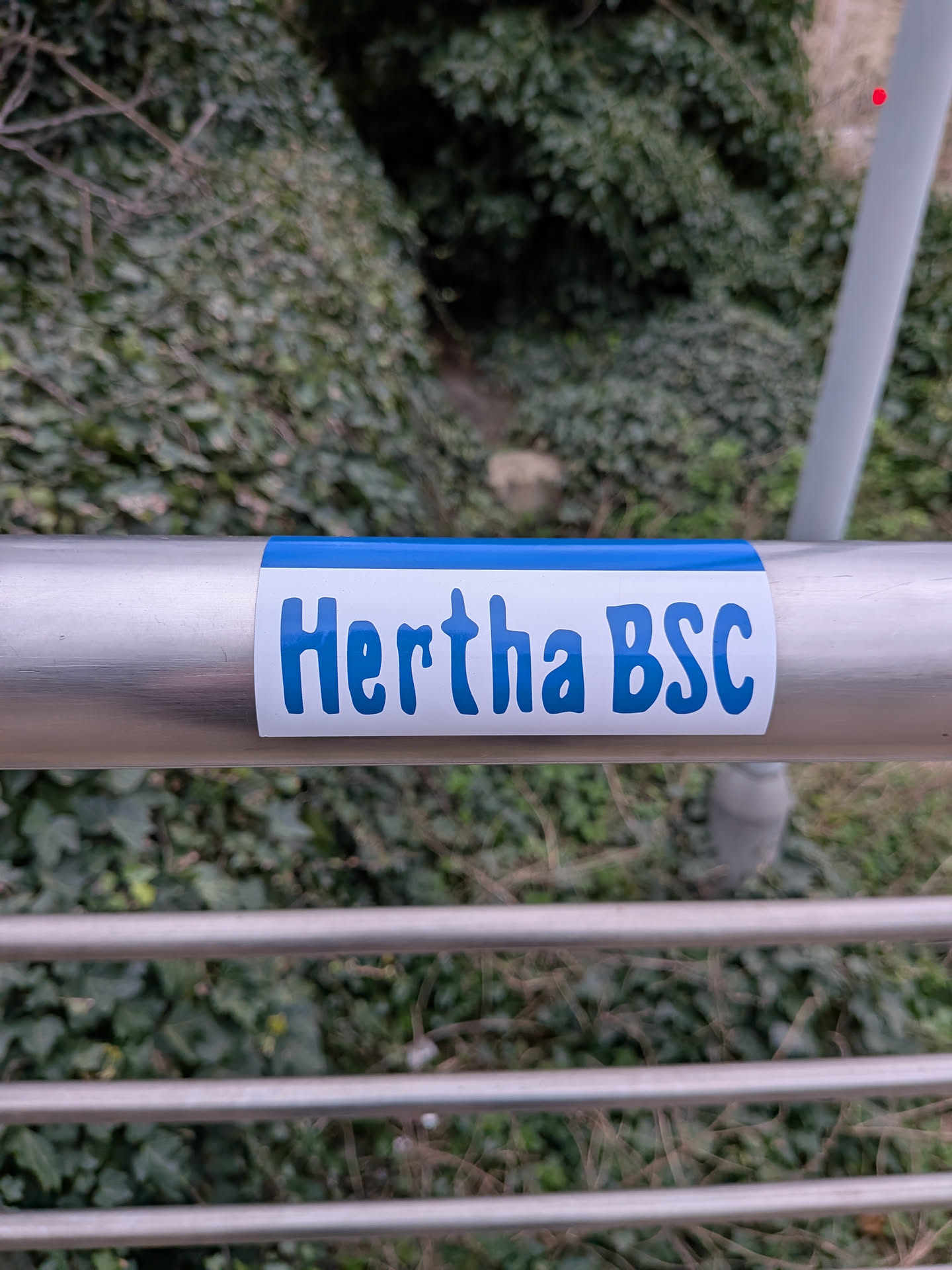 A rectangular sticker with slightly rounded corners, featuring a white background. A thin, solid blue strip runs along the top edge. The text "Hertha BSC" is prominently displayed in the center, rendered in a bold, slightly rounded, sans-serif font in a vibrant blue color. The sticker is affixed to a silver metal railing.