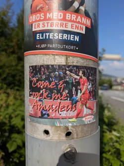 Street sticker The sticker prominently displays a football (soccer) player from SK Brann, dressed in a red uniform, in what appears to be a celebratory or action pose during a match, with a blurry stadium crowd in the background. The text 'Come & rock me Amadeus' is overlaid in a vibrant red script font, referencing the famous song by Falco. At the bottom right of the sticker, there's a small red oval with the word 'BRANN', clearly identifying the team. The sticker is affixed to a utility pole, with another larger poster above it also related to Brann, mentioning 'OBOS' and 'Eliteserien'.