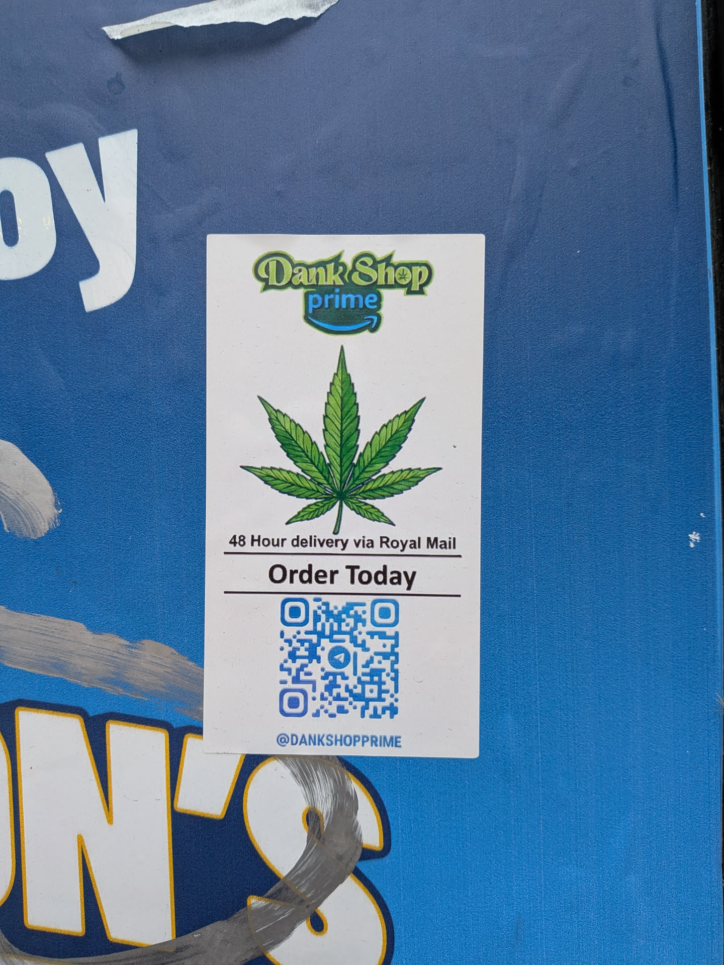 The sticker is a white rectangular label affixed to a textured blue surface. At the top, it features a logo that reads "Dank Shop" in a green, somewhat bubbly font, with "prime" underneath in a smaller blue font, styled similarly to the Amazon Prime logo, including a small blue arrow swoosh. Below the logo, a prominent, detailed illustration of a green cannabis leaf is centered. Further down, there are two lines of black text: "48 Hour delivery via Royal Mail" and "Order Today", with a horizontal line above and below "Order Today". The bottom half of the sticker contains a blue QR code. Below the QR code, a social media handle "@DANKSHOPPRIME" is printed in black text.