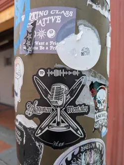 Street sticker A die-cut black sticker featuring a white illustration of a vintage microphone over crossed pens. It displays the text 'Lagunas Mentales' in gothic script, 'Alex' at the bottom, and a Spotify logo with a scannable code at the top.