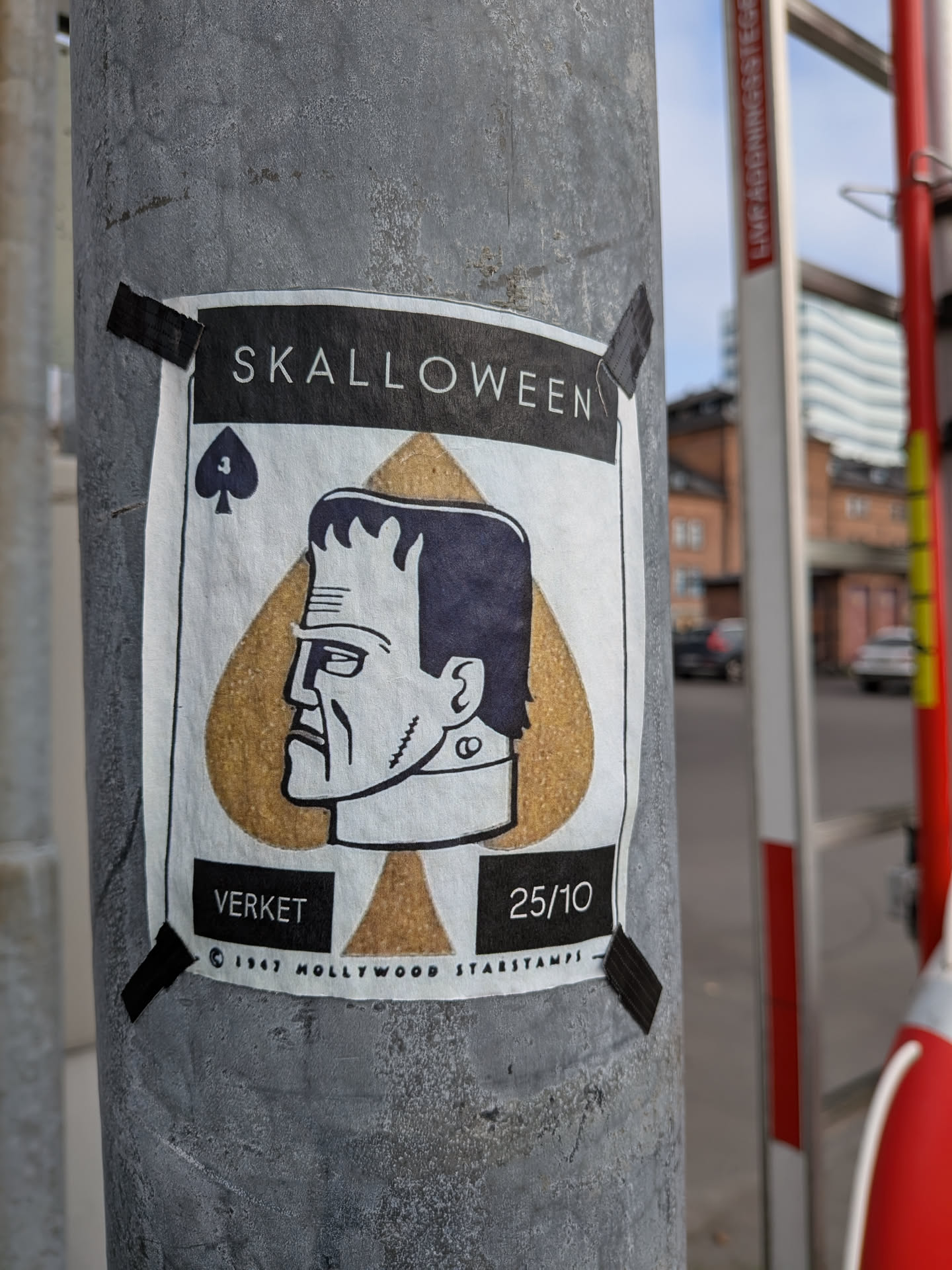 The sticker is centered on a grey pole and features a stylized depiction of Frankenstein's monster's head in profile. The monster's head is superimposed on a large, golden spade symbol, reminiscent of a playing card. Above the spade, the word 'SKALLOWEEN' is printed in white capital letters on a black background. Below the monster's head, there are two black rectangular boxes. The left box contains the word 'VERKET' in white text, and the right box contains '25/10'. At the very bottom, in small print, it reads '© 1947 HOLLYWOOD STARSTAMPS'. The sticker is attached to the pole with black tape in each corner.