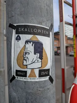 Street sticker The sticker is centered on a grey pole and features a stylized depiction of Frankenstein's monster's head in profile. The monster's head is superimposed on a large, golden spade symbol, reminiscent of a playing card. Above the spade, the word 'SKALLOWEEN' is printed in white capital letters on a black background. Below the monster's head, there are two black rectangular boxes. The left box contains the word 'VERKET' in white text, and the right box contains '25/10'. At the very bottom, in small print, it reads '© 1947 HOLLYWOOD STARSTAMPS'. The sticker is attached to the pole with black tape in each corner.