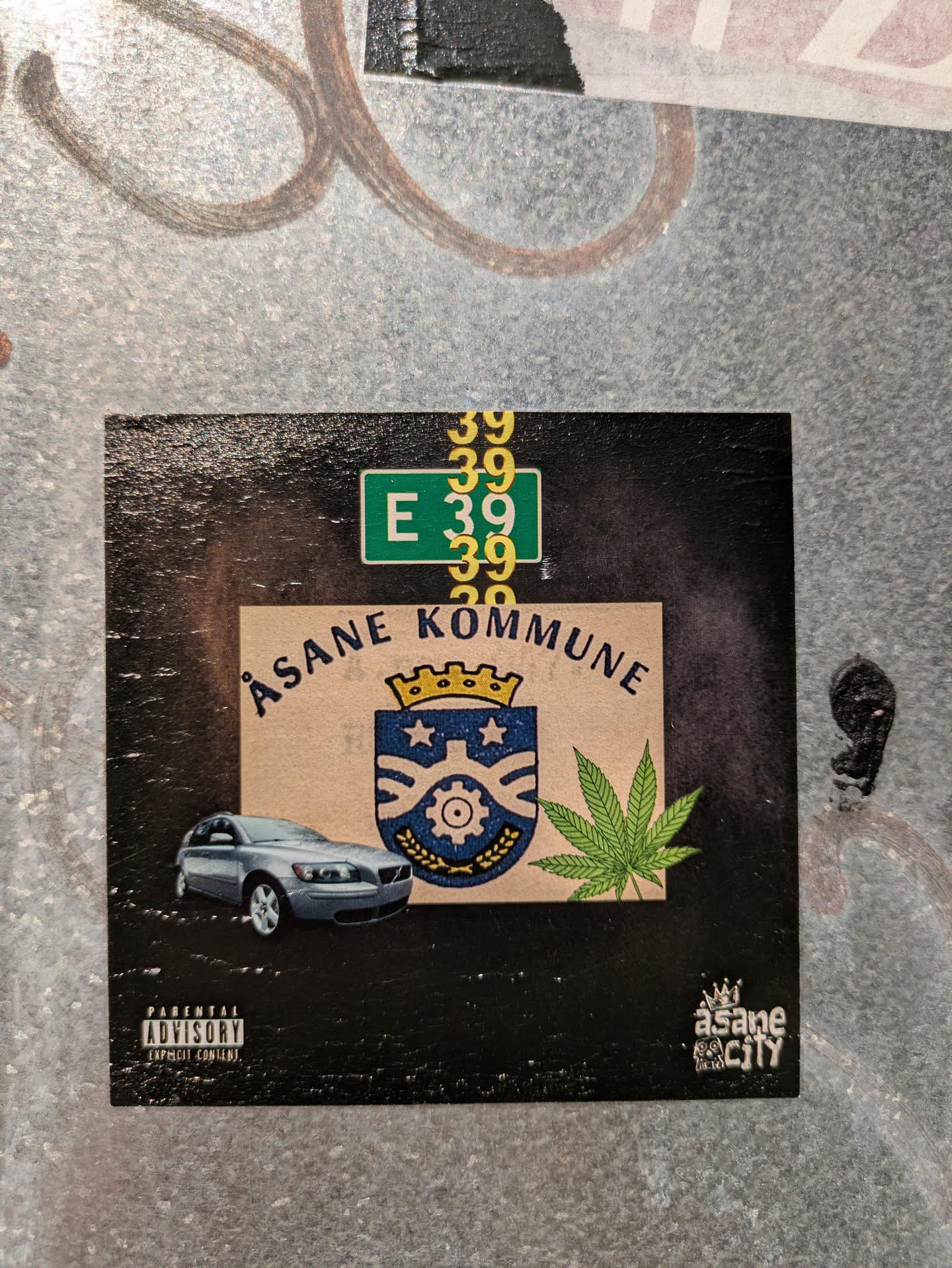 The sticker features a central beige-colored panel on a black background. At the top left, a green road sign displaying 'E 39' is visible, layered with multiple yellow '39' numbers. The beige panel presents the curved text 'ÅSANE KOMMUNE' above a blue and gold coat of arms with a crown, two stars, and a gear. To the left of the crest is a silver Volvo V50 station wagon, and to the right, a prominent green cannabis leaf. In the bottom left corner, the 'PARENTAL ADVISORY EXPLICIT CONTENT' logo is present. The bottom right corner contains a stylized white logo reading 'åsane CITY', topped with a small crown.