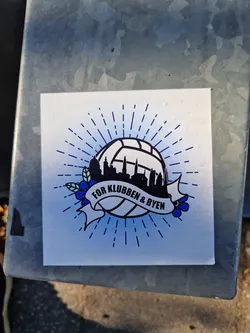 Street sticker A sticker featuring a stylized volleyball with a cityscape inside, surrounded by sunburst rays and floral accents.  The text on the banner reads "FOR KLUBBEN & BYEN".