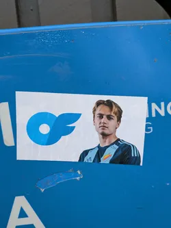Street sticker The sticker features a young man with blond hair, looking directly at the camera. He is wearing a dark blue football jersey with light blue stripes on the sleeves and a crest on the chest. To the left of the man is a stylized blue logo resembling a wing or a stylized 'f' with a circle in the center. The sticker is printed on white paper and appears to be adhered to a blue surface.