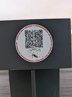 Round white sticker with a QR code in the center and a small illustration of a person falling down. The text around the QR code is barely legible, but it seems to be a repeated pattern. The sticker is attached to a dark gray surface.