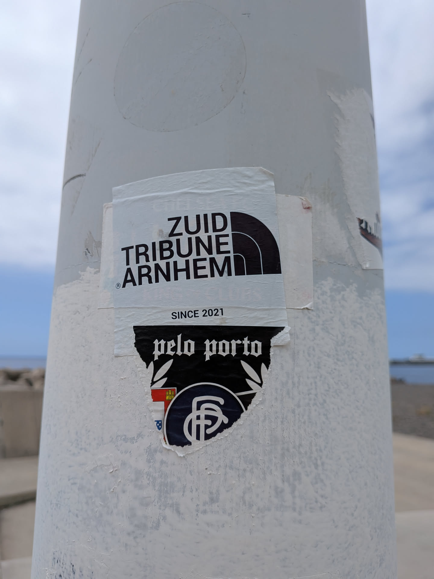 A white rectangular sticker featuring black text 'ZUID TRIBUNE ARNHEM' styled as a parody of The North Face logo, with 'SINCE 2021' printed below. It is pasted over a torn black sticker displaying 'pelo porto' and an FC Porto crest.