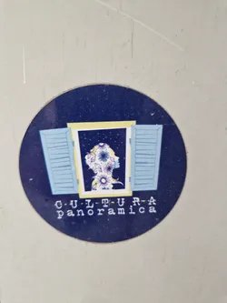 Round sticker featuring an illustration of a person's head and shoulders silhouetted against a night sky, framed by light blue shutters. The person's hair is depicted with purple flowers.  The words "CULTURA panoramica" are printed below the image.