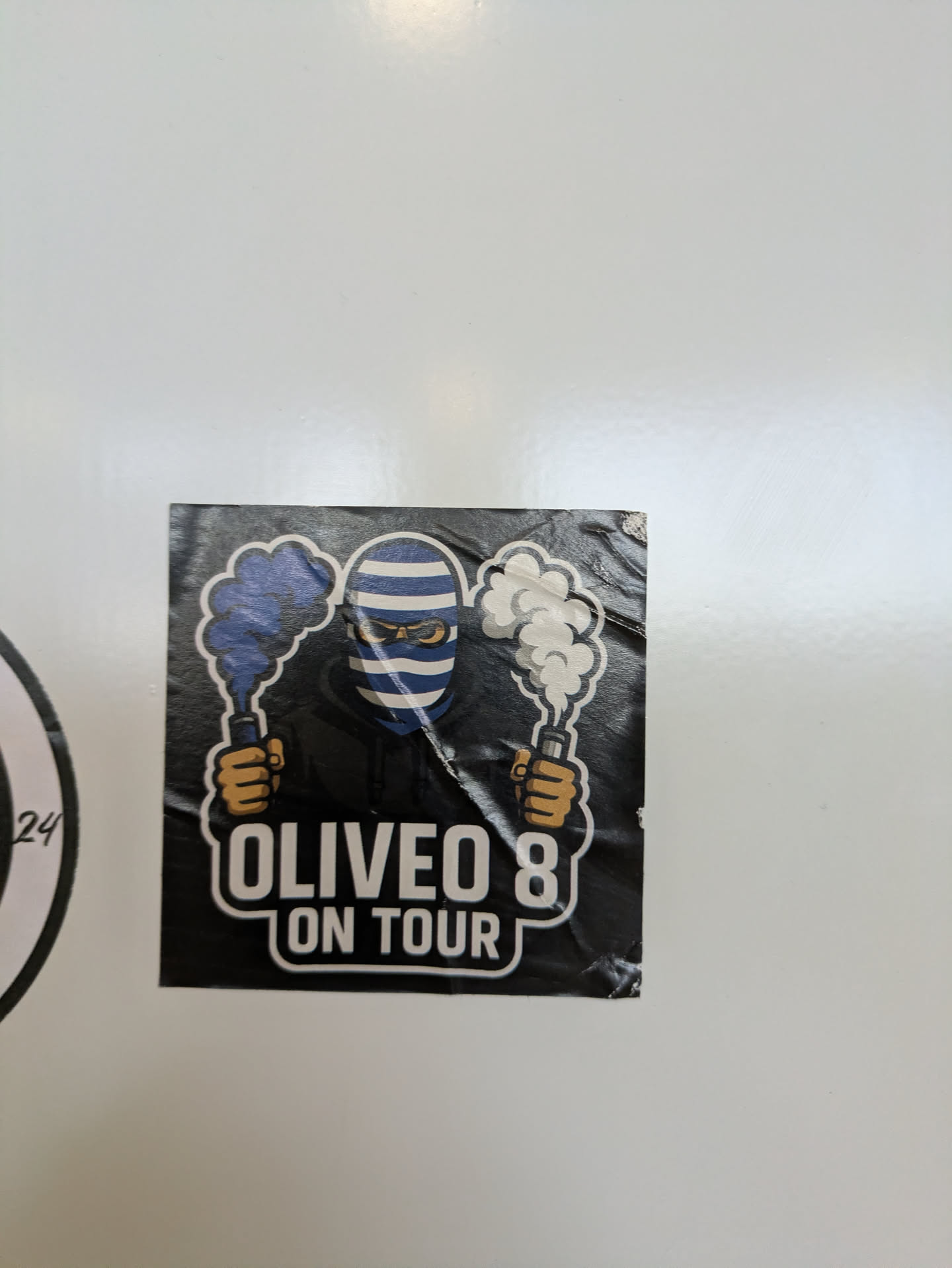 A square sticker with a black background. The image features a masked figure with blue and white striped face and yellow eyes, holding two flares that produce blue and white smoke. Below the figure, the text "OLIVEO 8 ON TOUR" is printed in a stylized font. The sticker appears to be placed on a white surface.