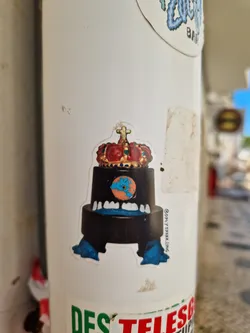 Street sticker A sticker featuring a cartoon monster wearing a crown. The monster is black with blue feet and has a face with large teeth. The crown is gold and red. The sticker is on a white background.