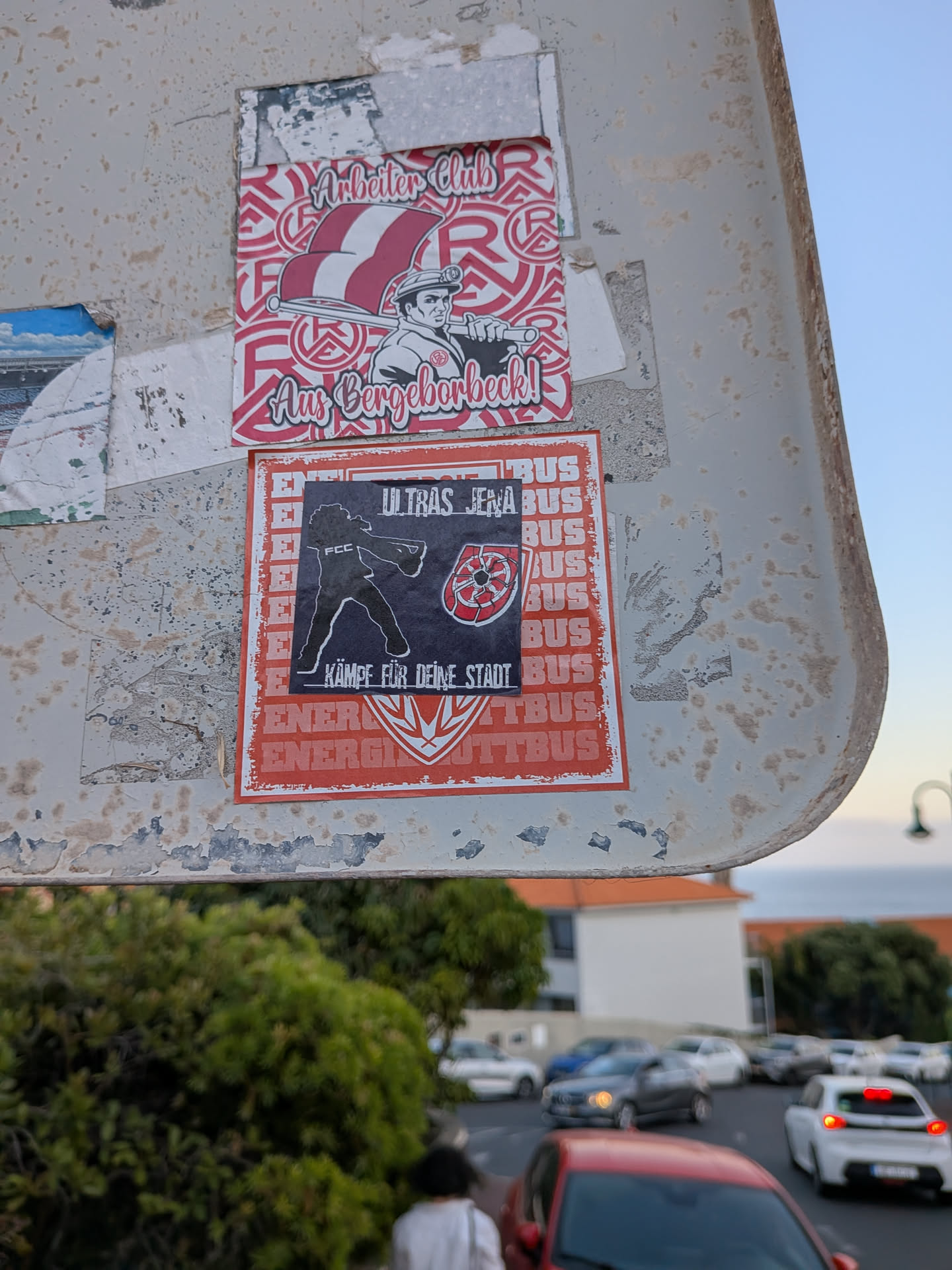 A black square sticker featuring a white silhouette of a supporter with 'FCC' on their back, a red and white cracked circular logo, and German text. It is layered on top of a larger red and white 'Energie Cottbus' sticker.