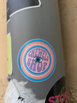 Street sticker The sticker features a circular design with concentric rings of light blue, pink, and white. In the center, the text "CHEWELS SHOP" is written in a wavy, psychedelic font in shades of purple and pink. An eye with long eyelashes and a blue iris is placed between the two words. The overall style evokes a vintage, groovy aesthetic.
