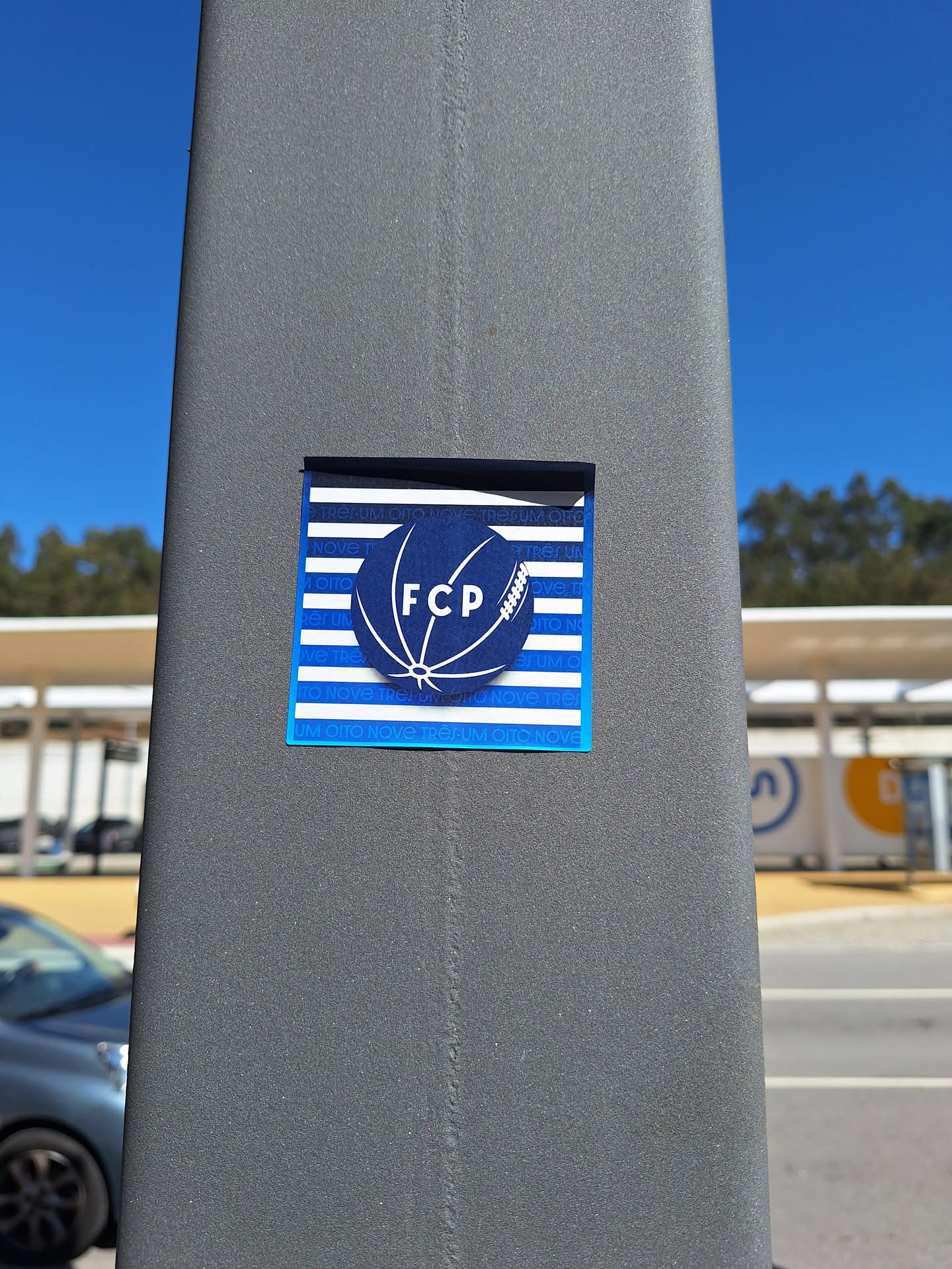 A rectangular sticker with a dark blue border, affixed to a textured grey pole. The background features alternating horizontal blue and white stripes. Superimposed on the blue stripes, the text 'UM OITO NOVE TRÊS' is subtly repeated in a lighter blue. At the center, a large dark blue circle contains a white outline of a stylized ball, resembling a hybrid of a soccer and rugby ball. Inside this ball, the letters 'FCP' are prominently displayed in white capital letters.