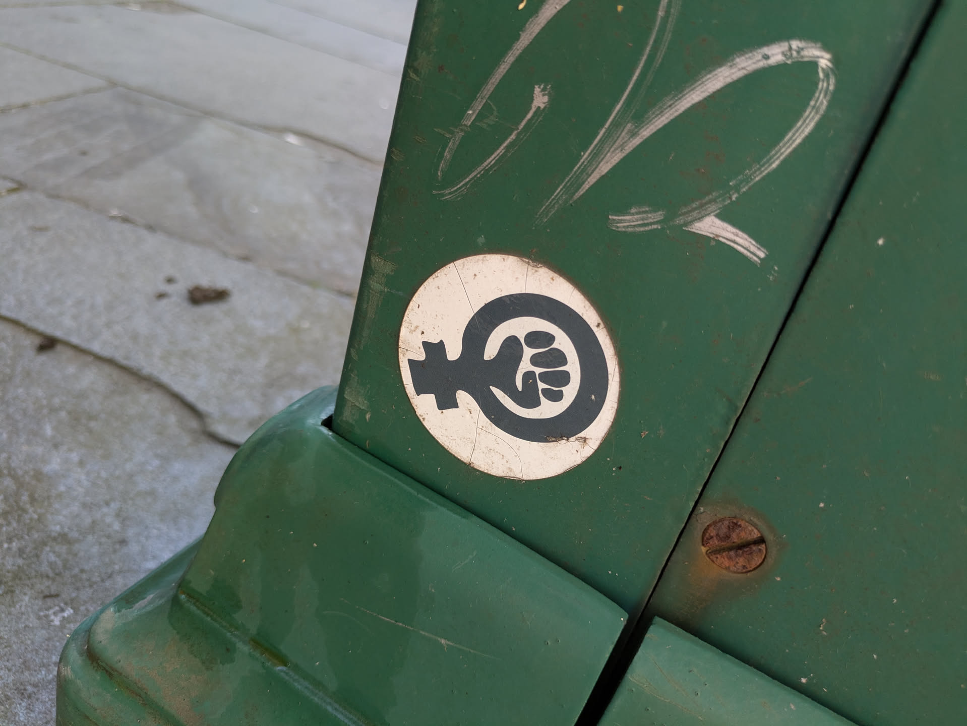 A circular sticker adheres to a weathered, dark green metal surface, likely a utility box or cabinet. The sticker has a white background with a prominent black graphic. This graphic ingeniously combines the traditional female gender symbol (Venus symbol) with a clenched, raised fist integrated into the loop of the symbol, symbolizing female empowerment, solidarity, and resistance. The sticker shows slight signs of wear, including minor cracks on its surface, indicating exposure to the elements. Above the sticker, there are visible white scratch marks on the green surface, and a rusty screw is located to the right.