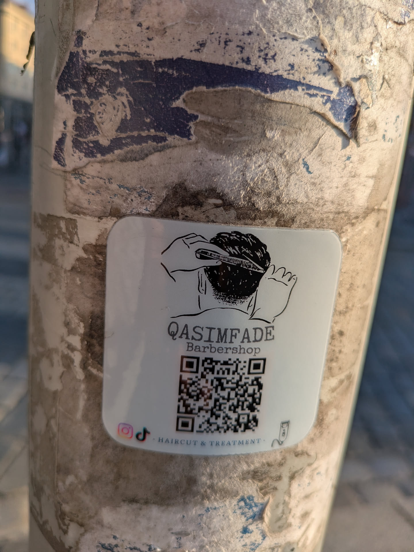A white, square-shaped sticker with rounded corners is affixed to a weathered, peeling pole. The sticker prominently displays a black line-art illustration of a person's head being given a haircut with clippers and a comb. Below this illustration, the text 'QASIMFADE Barbershop' is printed. A large, scannable QR code occupies the central lower portion of the sticker. Below the QR code, the phrase 'HAIRCUT & TREATMENT' is visible. To the left of this text, there are small logos for Instagram and TikTok, indicating their social media presence.