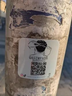 Street sticker A white, square-shaped sticker with rounded corners is affixed to a weathered, peeling pole. The sticker prominently displays a black line-art illustration of a person's head being given a haircut with clippers and a comb. Below this illustration, the text 'QASIMFADE Barbershop' is printed. A large, scannable QR code occupies the central lower portion of the sticker. Below the QR code, the phrase 'HAIRCUT & TREATMENT' is visible. To the left of this text, there are small logos for Instagram and TikTok, indicating their social media presence.