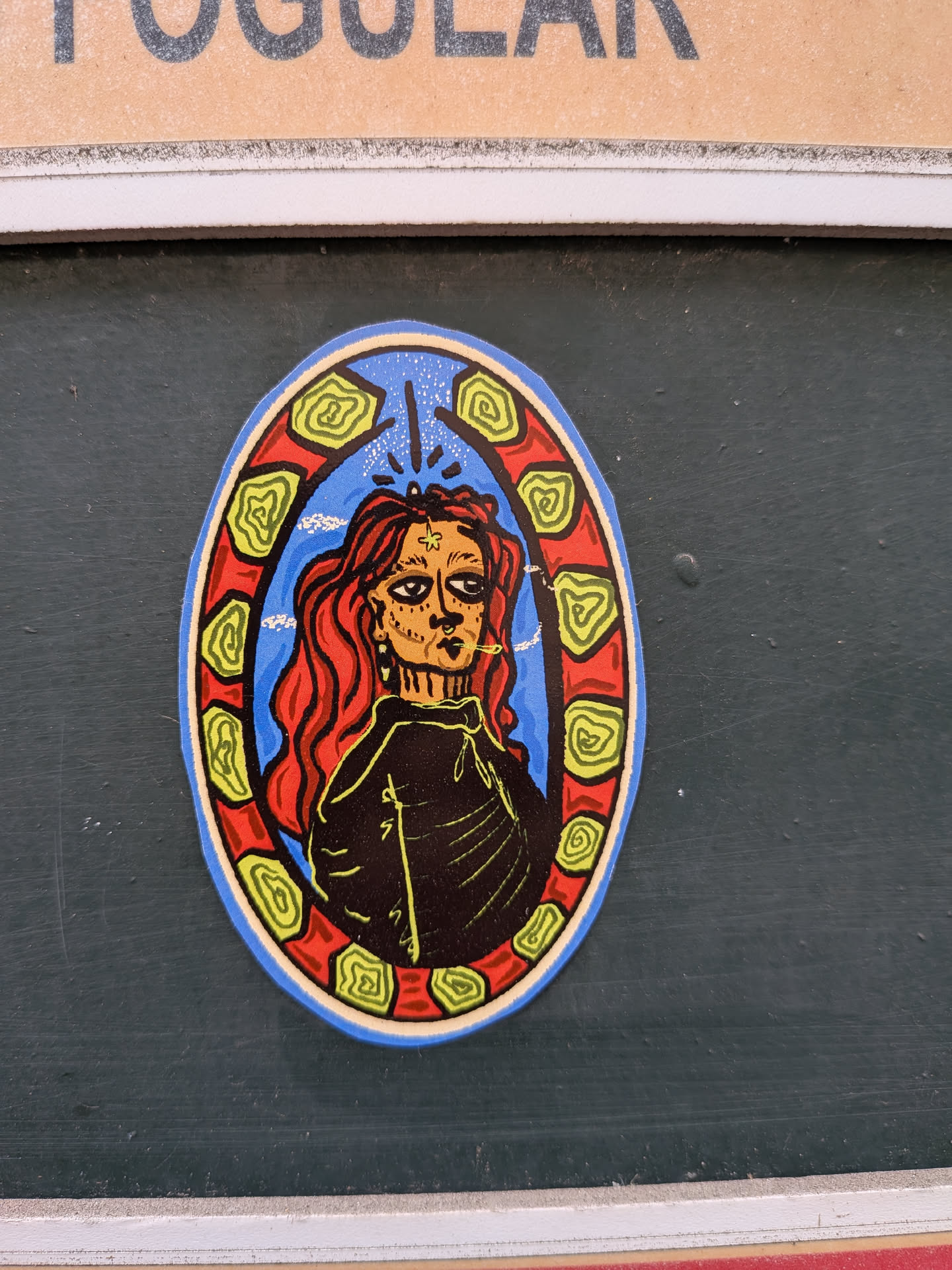 An oval sticker depicting a stylized portrait of a person with long red hair and a star on their forehead, smoking a cigarette. The figure is set against a blue sky background and framed by a red border decorated with green spiral patterns.