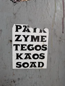 A rectangular white sticker with bold black text in a stylized serif font, pasted on a weathered grey surface. The text lists five names vertically: PAIK, ZYME, TEGOS, KAOS, and SOAD. The top edge of the sticker is torn and damaged.