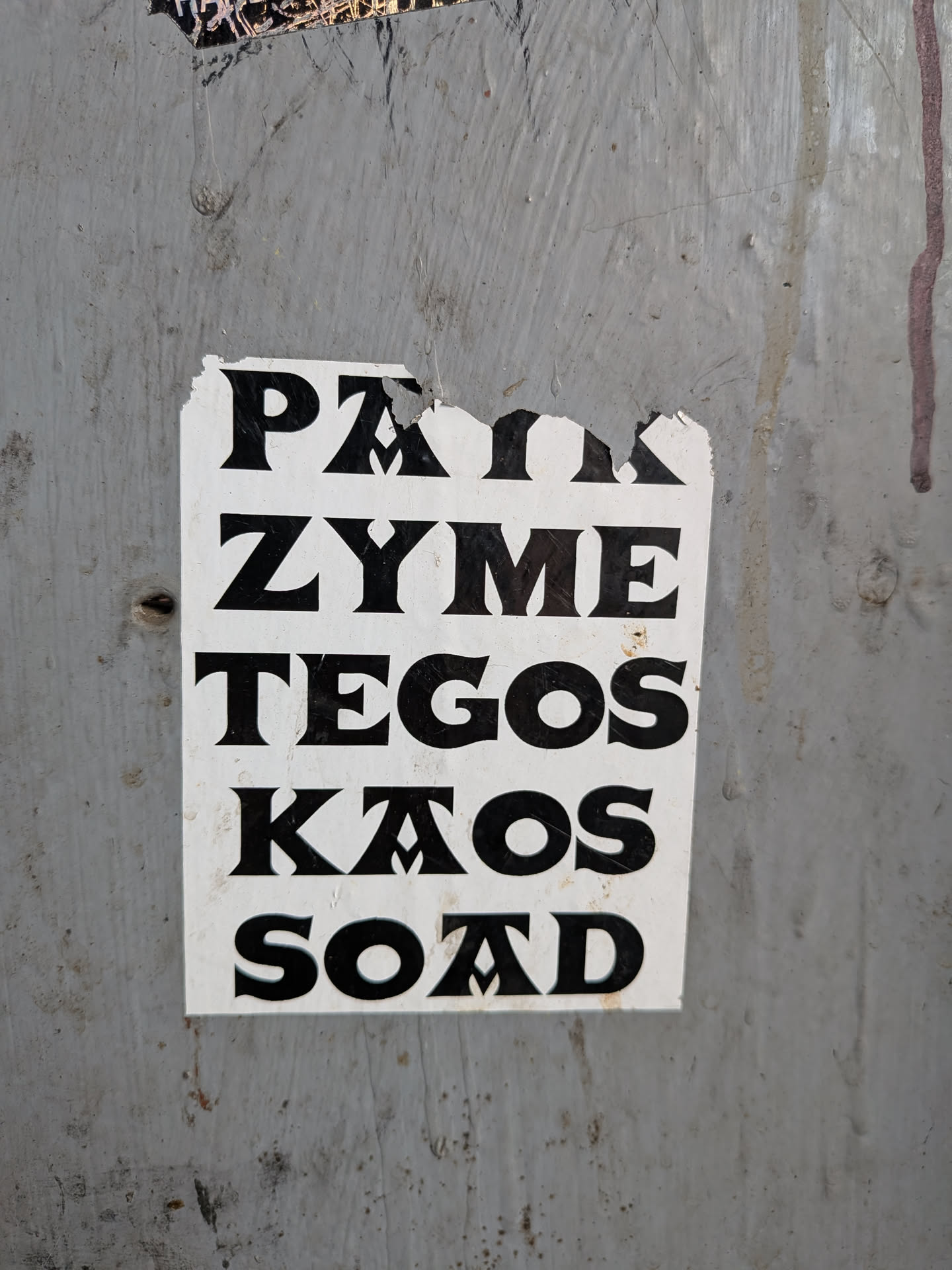 A rectangular white sticker with bold black text in a stylized serif font, pasted on a weathered grey surface. The text lists five names vertically: PAIK, ZYME, TEGOS, KAOS, and SOAD. The top edge of the sticker is torn and damaged.