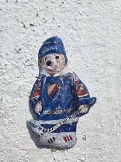 Street sticker The sticker depicts a cartoonish bear wearing a blue hockey uniform. The bear has a light brown face with black eyes and nose, and a small smile. It wears a blue beanie with white lettering that is partially obscured. Its jersey is predominantly blue with red and white accents, featuring the team logo on the chest, which appears to be a shield with red and gold elements and possibly a crown. The jersey also has the number '22' on the sleeves. The bear is holding a hockey stick, and its feet are depicted in hockey skates with red Adidas stripes on the sides. The overall style is somewhat naive and hand-drawn, with visible texture suggesting it's a physical sticker applied to a rough, white surface.
