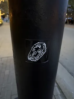 Street sticker The sticker is rectangular with slightly rounded corners, featuring a white illustration on a solid black background. The main image is a stylized, somewhat crudely drawn coffin, depicted from a slightly elevated perspective. Inside the coffin lies a white, ghost-like or skeletal figure with large, hollow eyes and a wavy, melting body that forms a W-like shape. Along the bottom edge of the coffin, the word 'GLASS' is written in a blocky, hand-drawn font. On the right side of the coffin, running vertically, is the word 'COFFIN' in a similar style. A small, stylized 'W' is visible on the upper right side of the coffin's exterior. The overall aesthetic is stark, monochrome, and somewhat macabre.