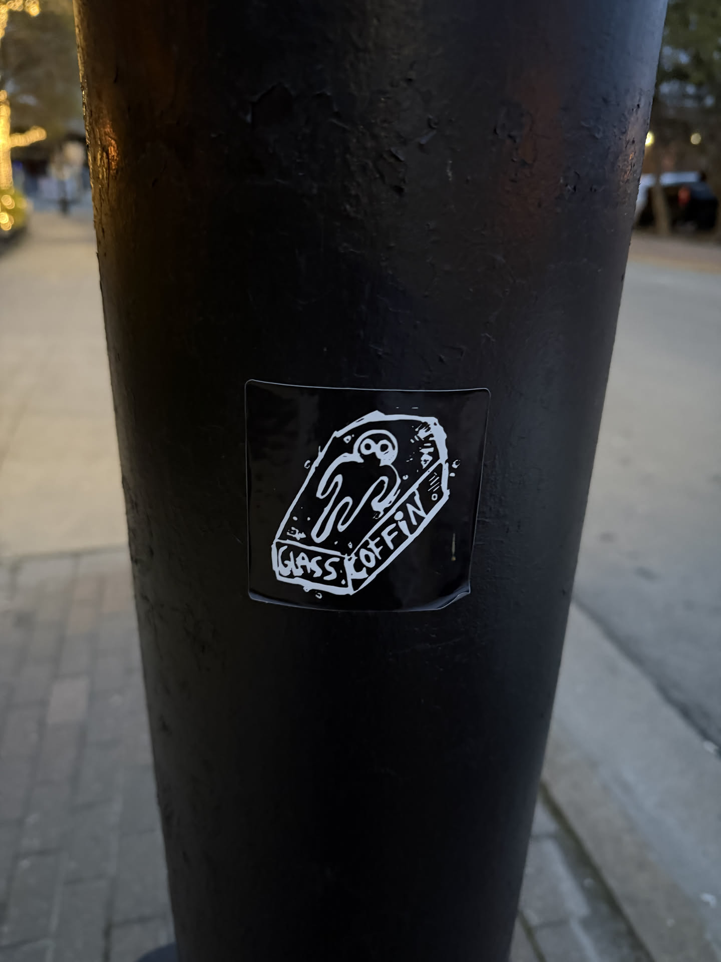 The sticker is rectangular with slightly rounded corners, featuring a white illustration on a solid black background. The main image is a stylized, somewhat crudely drawn coffin, depicted from a slightly elevated perspective. Inside the coffin lies a white, ghost-like or skeletal figure with large, hollow eyes and a wavy, melting body that forms a W-like shape. Along the bottom edge of the coffin, the word 'GLASS' is written in a blocky, hand-drawn font. On the right side of the coffin, running vertically, is the word 'COFFIN' in a similar style. A small, stylized 'W' is visible on the upper right side of the coffin's exterior. The overall aesthetic is stark, monochrome, and somewhat macabre.