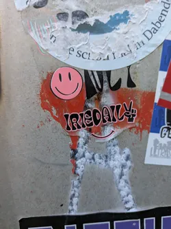Street sticker The central sticker prominently features the brand name "IRIEDAILY" in bold black letters with a distinct pink outline. The letter 'Y' in "IRIEDAILY" is cleverly replaced by a black peace sign symbol, also with a pink outline. Above this main text, a classic pink smiley face with black eyes and mouth is displayed. Below the "IRIEDAILY" text, a minimalist pink smiley mouth line can be observed. The sticker is applied on a light gray concrete surface, which is heavily marked with vibrant red spray paint splatters and some white, possibly paint or residue, giving it a raw, urban feel.