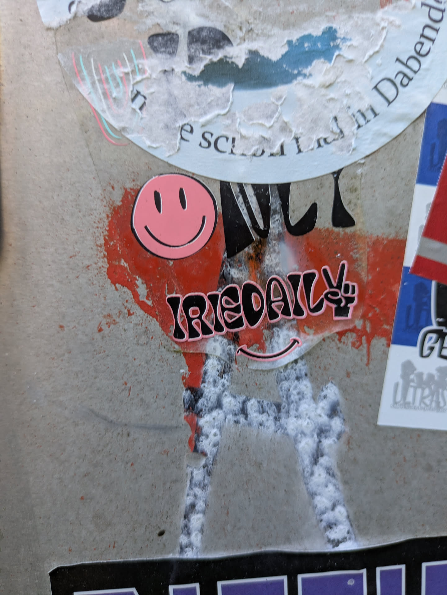 The central sticker prominently features the brand name "IRIEDAILY" in bold black letters with a distinct pink outline. The letter 'Y' in "IRIEDAILY" is cleverly replaced by a black peace sign symbol, also with a pink outline. Above this main text, a classic pink smiley face with black eyes and mouth is displayed. Below the "IRIEDAILY" text, a minimalist pink smiley mouth line can be observed. The sticker is applied on a light gray concrete surface, which is heavily marked with vibrant red spray paint splatters and some white, possibly paint or residue, giving it a raw, urban feel.