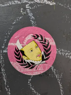 This round sticker has a pink background and shows a portrait of a person with light yellow skin. They are wearing a white ruffled collar and a pink hat with the word "KAMIONEY" written on it. A laurel wreath in black surrounds the portrait.