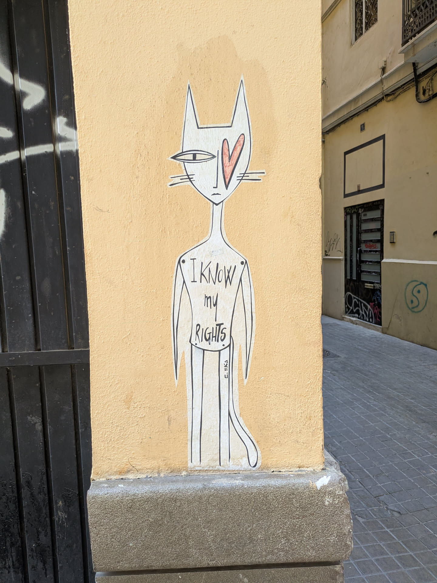 A white, stylized cat sticker affixed to a yellow wall. The cat has a slender body, thin limbs, and large pointed ears. One of its eyes is a solid red heart. Text on the body reads "I know my rights" in all caps. The signature "C.SKA" is visible at the bottom right of the sticker.