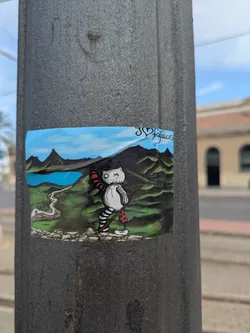The sticker features a cat-like character with a striped body and red-and-black arm warmer and knee pad, holding a walking stick. It stands in front of a painted landscape with a lake, mountains, and a blue sky. The artist's name is written at the top right corner of the landscape: "3 corações falalas".