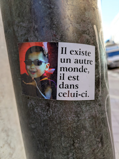 A sticker featuring a child wearing sunglasses next to text in French that reads: "Il existe un autre monde, il est dans celui-ci.", which translates to "There is another world, it is in this one."