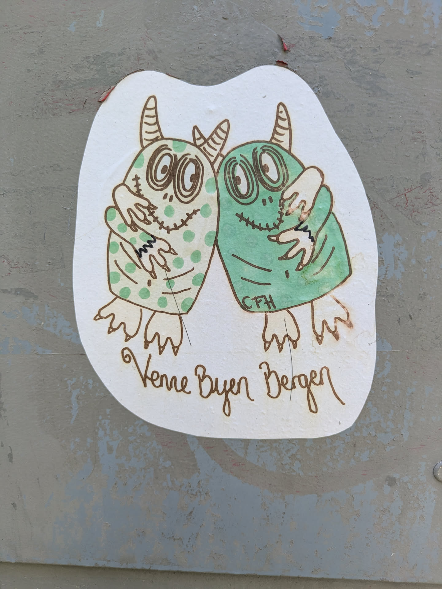 The sticker depicts two cartoon-style, green monsters hugging. The monster on the left is a lighter green with darker green polka dots, while the monster on the right is a solid medium green. Both creatures have large, expressive eyes with concentric circles, stitched mouths resembling stitches, and small horns on their heads. They embrace each other with three-fingered hands. Below the monsters, the phrase "Venner Byen Bergen" is written in a brown, flowing script. Small initials "CFH" are also visible on the lower part of the right monster's body. The sticker itself is cut out in an irregular shape and placed on a weathered, light grey surface.