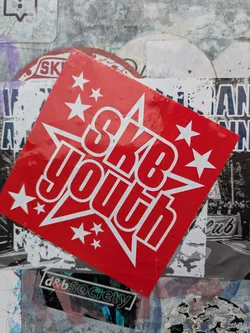 Street sticker The central sticker is a bright red square, rotated diagonally to resemble a diamond. Prominently displayed in white is the text "SKB youth", with 'SKB' above 'youth'. The text is encased within a large white star outline, and several smaller white stars are scattered around the main text, creating a dynamic, celebratory feel. The sticker appears to be layered over older, worn stickers and textured surfaces, suggesting it's part of a larger sticker bomb or collection on an urban surface.