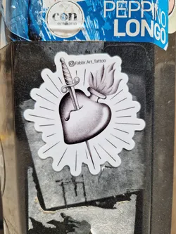 Street sticker Black and white sticker with a design of a sacred heart pierced by a dagger with a flame on top. The sticker has a sunburst effect around it and the artist's Instagram handle is mentioned on it.