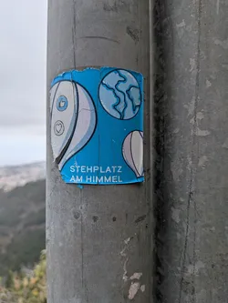 Street sticker A weathered blue sticker on a metal pole. It features illustrations of hot air balloons with social media icons, a globe, and the German text 'STEHPLATZ AM HIMMEL'.