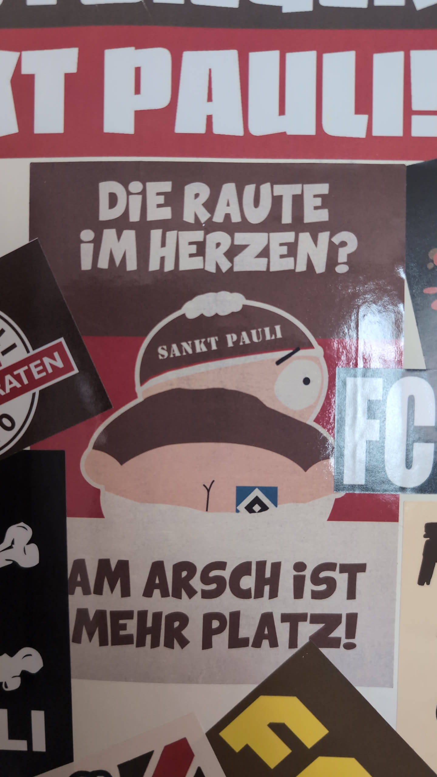 The sticker features a cartoon character, strongly resembling Eric Cartman from South Park, depicted from behind, bending over. The character wears a brown hat with 'SANKT PAULI' written in white. On the character's exposed buttocks, there is a small blue and white diamond logo, which is the emblem of Hamburger SV (HSV). The background of the sticker is brown. Above the character, the text 'DIE RAUTE IM HERZEN?' is written in white. Below the character, the text 'AM ARSCH IST MEHR PLATZ!' is written in white. The overall color scheme is brown, white, and skin tone, with a small blue and white accent for the HSV logo. The style is cartoonish and provocative, clearly referencing the football rivalry between FC St. Pauli and HSV.