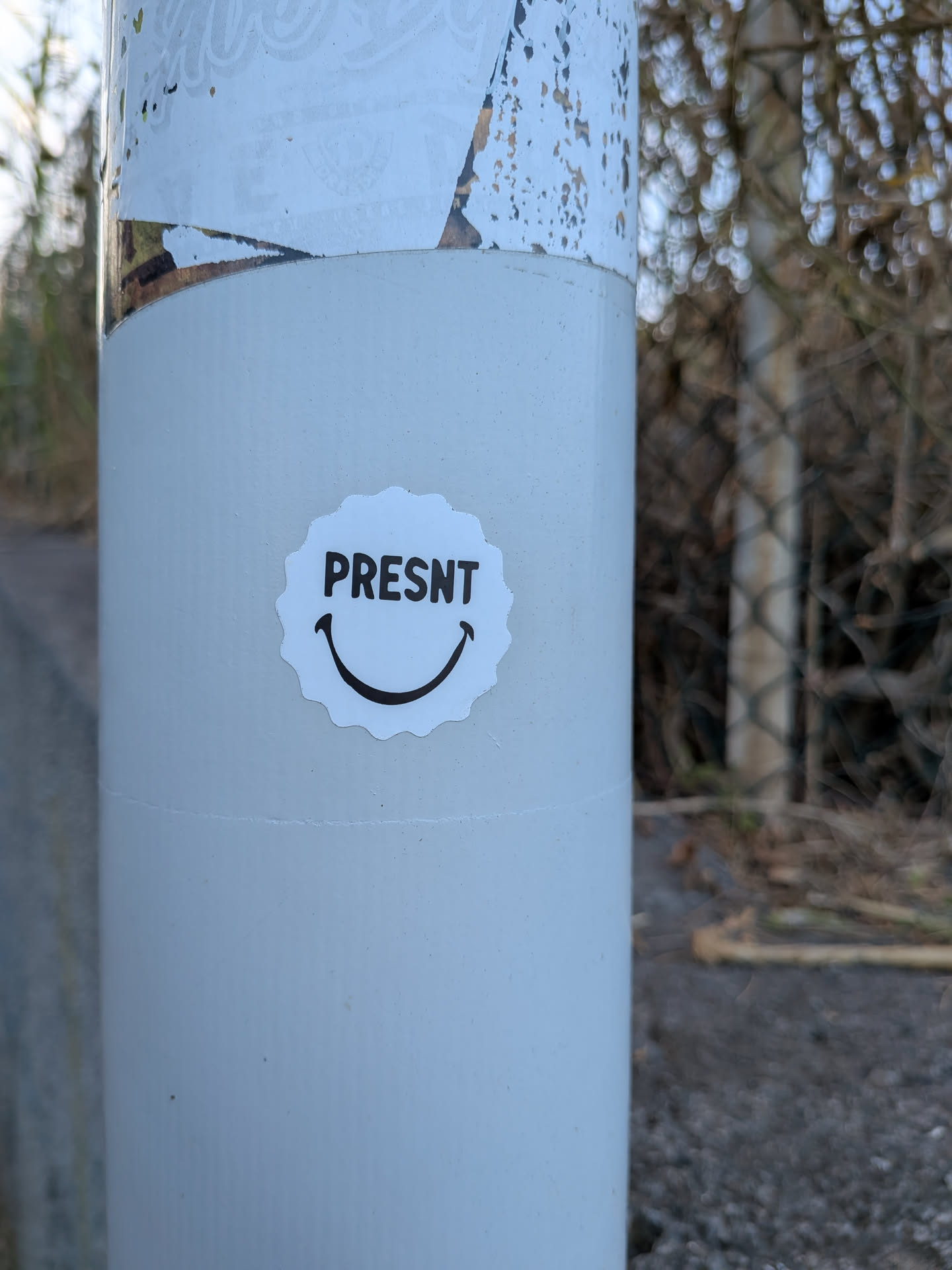 A scalloped-edge white sticker on a grey pole, featuring the black text 'PRESNT' above a simple curved smile.