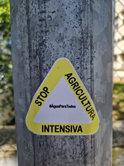Street sticker Triangular yellow sticker with the text "STOP AGRICULTURA INTENSIVA" and the hashtag #AguaParaTodos in the center. The sticker is attached to a gray metal pole.