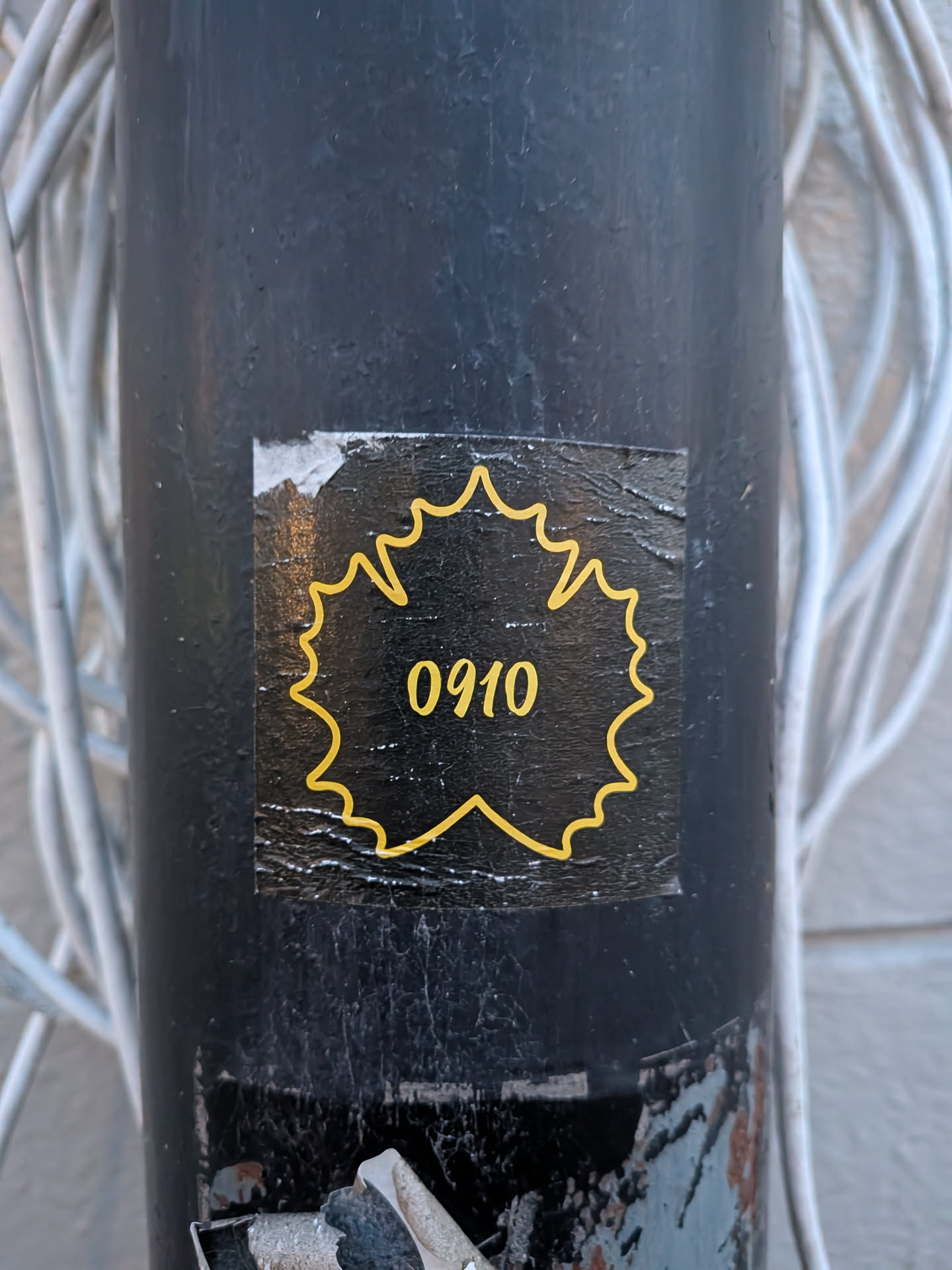 The sticker is predominantly black with a vibrant yellow outline of a stylized grape leaf. Inside the leaf shape, the numbers '0910' are displayed in the same yellow color. The sticker appears to be slightly worn and adhered to a dark, cylindrical surface, possibly a pole, with some visible texture and minor peeling at the edges.