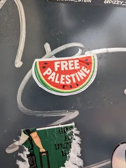 A semi-circular sticker designed as a watermelon slice with a red center, black seeds, and a green rind. The bold white text 'FREE PALESTINE' is printed across the red section.
