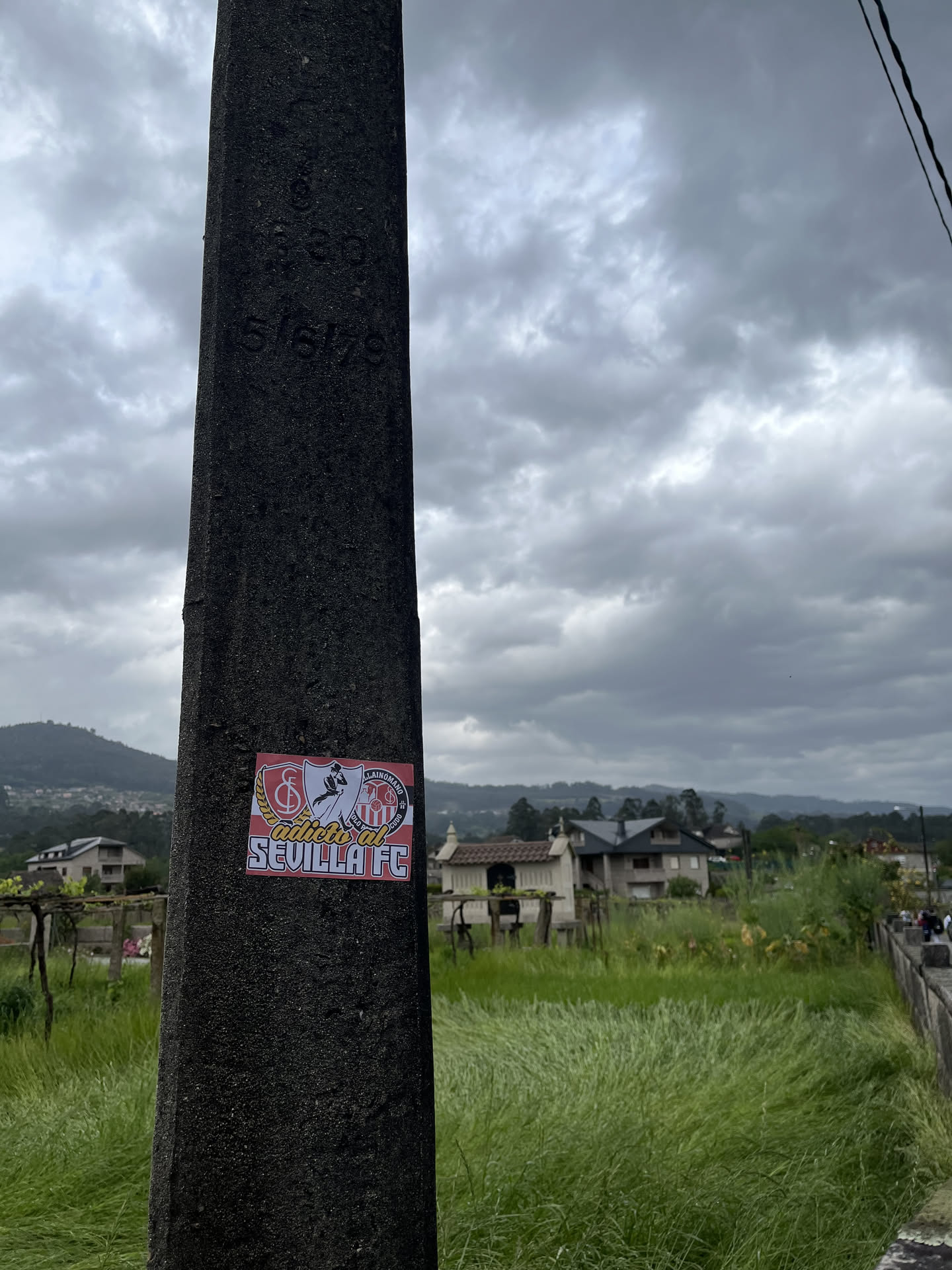 A rectangular sticker with a prominent red border, affixed to a dark, textured concrete pole. The design features a collage of elements associated with Sevilla FC. On the left, a gold laurel wreath encircles a crest with the letters 'SFC' in white on a red background. In the center, a black silhouette of a figure, possibly a bullfighter or a traditional dancer, is depicted. To the right, another shield-like crest with vertical red and white stripes is visible, topped by smaller text including 'LLAIMOMANO' and 'VIDA'. The bottom section of the sticker has a red background with the white text 'adictos al SEVILLA FC' in a bold, sans-serif font. The overall color scheme is red, white, black, and gold.