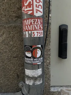 Street sticker A rectangular sticker featuring a stylized cartoon character on the left and a circular football club logo on the right, set against a predominantly black background. The character wears a black bucket hat, yellow-rimmed glasses, and a white face mask, with a stern expression. The circular logo is split vertically into red and white halves, with a large white 'F' in the center. The words 'FEYENOORD' and 'ROTTERDAM' are written in gold/yellow text around the 'F' within a black outer ring. Below the character, the text 'NERGENS WELKOM' is in white, and below the logo, 'OVERAL AANWEZIG' is also in white, both on a black background.